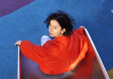Singer E^ST wears a red shirt and sits on a slide while she looks back at the camera