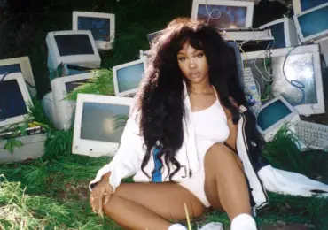Artist, SZA, sits down in a white bodysuit and matching jacket and shoes in front of computer monitors set up aesthetically.